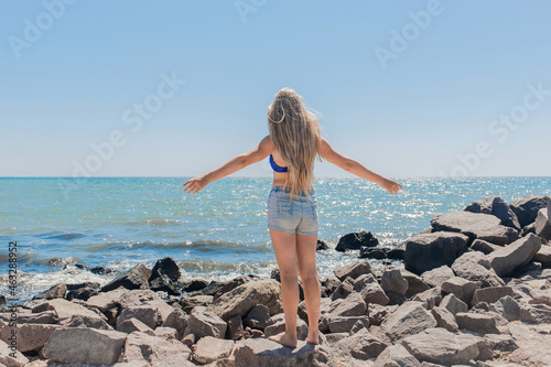 A young, teenage girl with long hair blonde stands on the rocks near the sea shore on the beach spreading and raising her hands to the sides