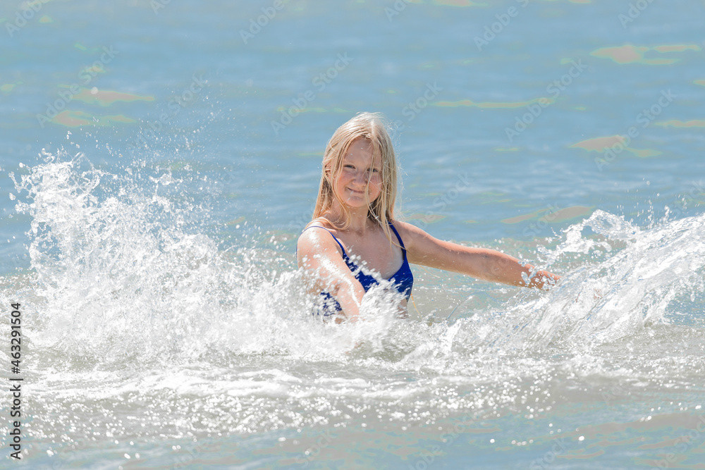 Young cheerful girl teenager blonde European appearance makes splashes of water with her hands in the sea