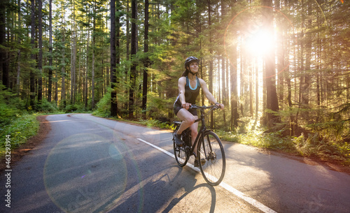 Fototapeta Naklejka Na Ścianę i Meble -  Adventurous White Caucasian Woman on a bicycle riding on a path in Green Canadian Rain Forest. Sunny Sunset. Seymour Valley Trailway in North Vancouver, British Columbia, Canada.