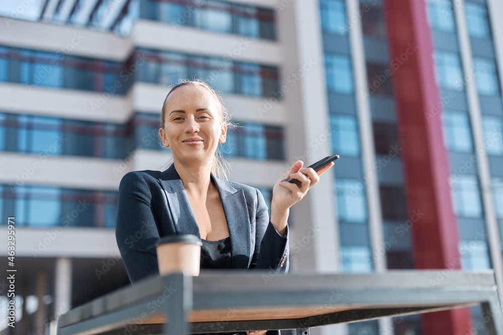 Smiling european business woman has coffee break, stands outside of office by table, drinks coffee from takeaway cup, holds phone in hand, wears ponytail, business suit and deep neckline blouse.