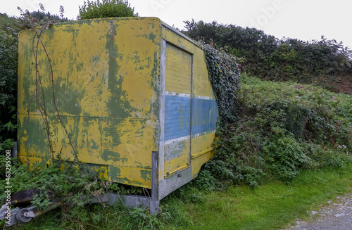 Abandoned weathered colourful industrial trailer, overgrown with ivy left by a roadside. Towing hitch Flaking yellow, blue and white paintwork. Landscape image with space for text. UK. © Steve