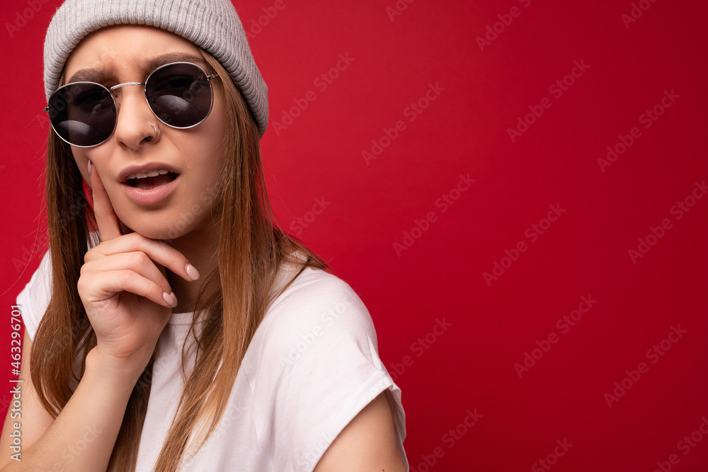 Fototapeta premium Closeup of young dissatisfied thoughtful emotional beautiful dark blonde woman with sincere emotions wearing casual white t-shirt, gray hat and sunglasses isolated on red background with copy space