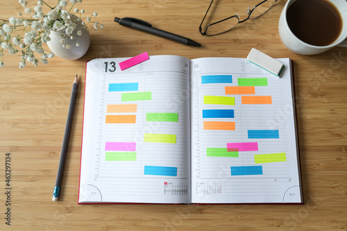 Open diary or appointment calendar with a lot of colorful sticky notes on a wooden desk with coffee pens and glasses, time management concept, copy space, selected focus, flat lay photo