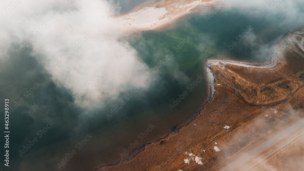 Aerial drone view from above of a dune beach shore line with morning sunlight and foggy clouds over the ocean. Beautiful turquoise green north sea water with sand bankes in the river. Denmark