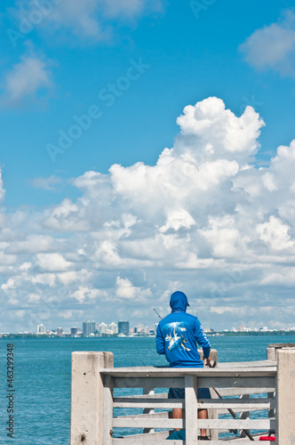 back view, ar distance of a middle aged, Mal with a blue ball cap, fish off a wood dock, on a tropical island on a clear , sunny day