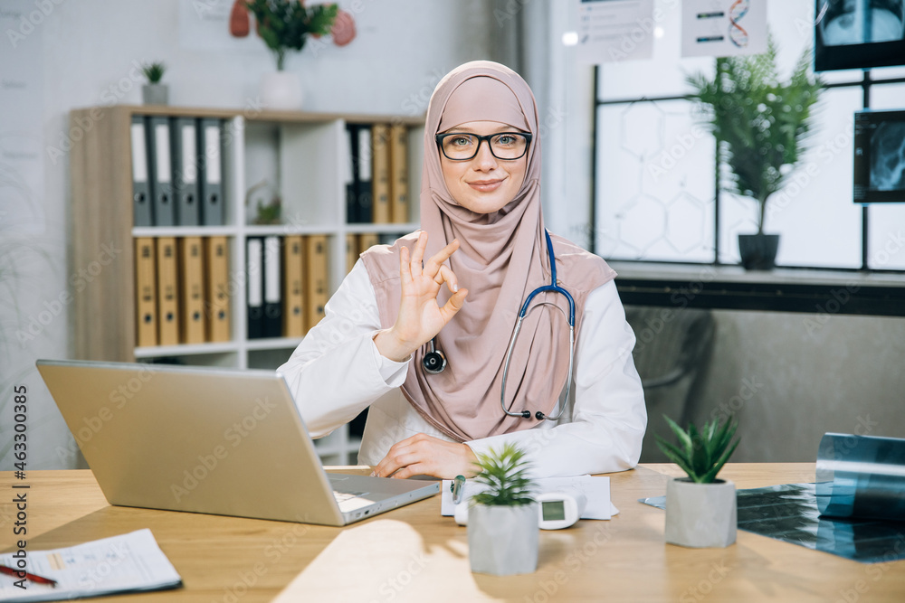 Portrait of beautiful arabian woman in hijab and eyewear sitting at desk with modern laptop and showing okay approval gesture at camera. Qualified female doctor having online work Portrait of beautiful arabian woman in hijab and eyewear sitting at desk with modern laptop and showing okay approval gesture at camera. Qualified female doctor having online work