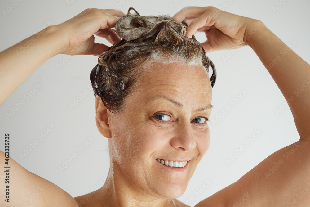 Naklejka premium Mature woman with bare shoulders washing hair with shampoo, smiling looking at camera
