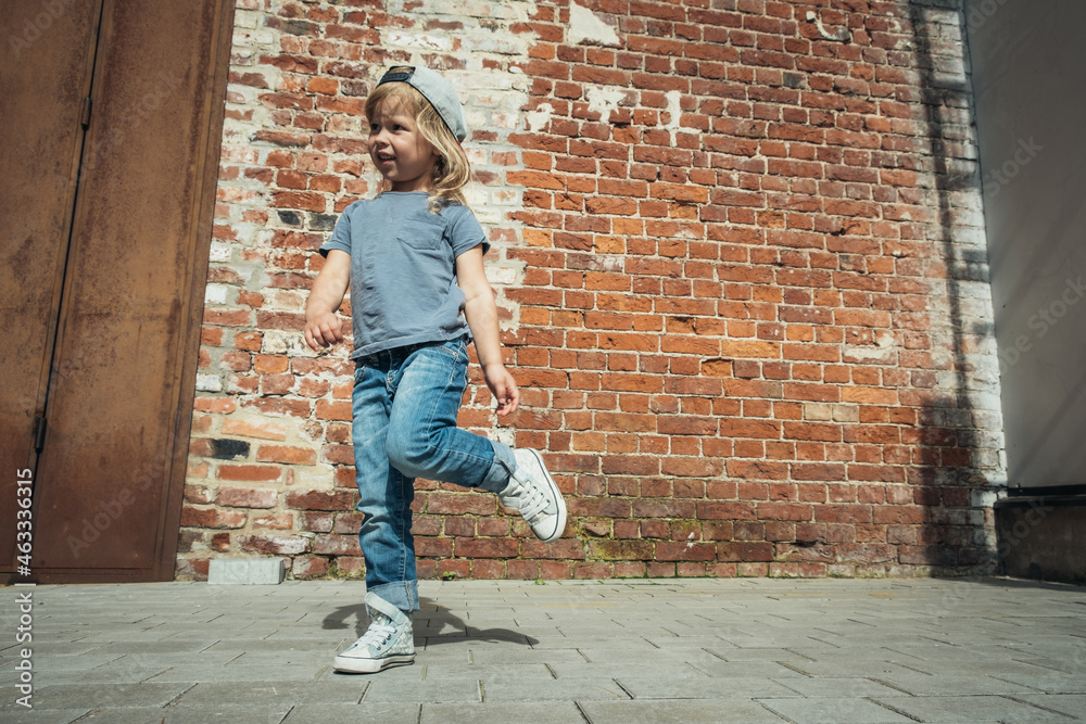Little cute girl in a blue t-shirt and jeans with a cap is dancing on the background of an old brick wall. Little cute girl in a blue t-shirt and jeans with a cap is dancing on the background of an old brick wall.
