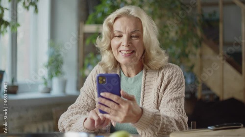 A woman in a sweater and with gray hair is talking on a video call using a phone. She talks sweetly and smiles a lot