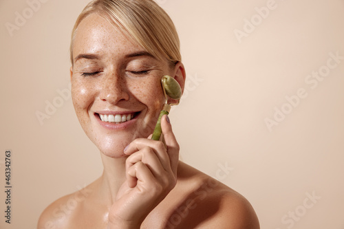 Smiling woman with freckles using jade stone roller against pastel background