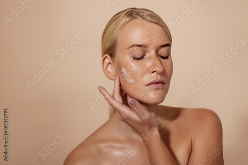 Caucasian woman applying facial moisturizer standing in studio against pastel background