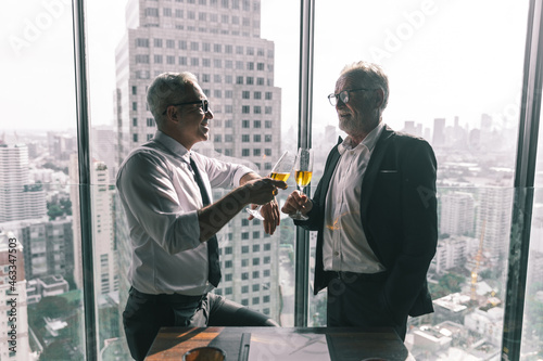 Old business man and young business man drinking champagne to celebrate their success. They are standing by the window in a hotel lobby with beautiful cityscape view behind.