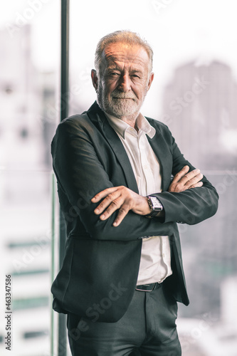 Old business man portrait. He is lookin into the camera with his arm wrap around his body. He is in black suit with white shirt.