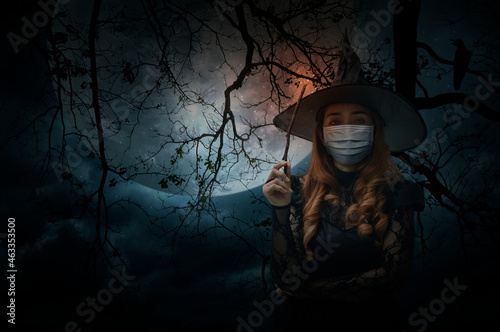 Halloween witch wearing medical face mask holding magic wand standing over dead tree, full moon, bird and spooky cloudy sky, Halloween and coronavirus or covid-19 concept