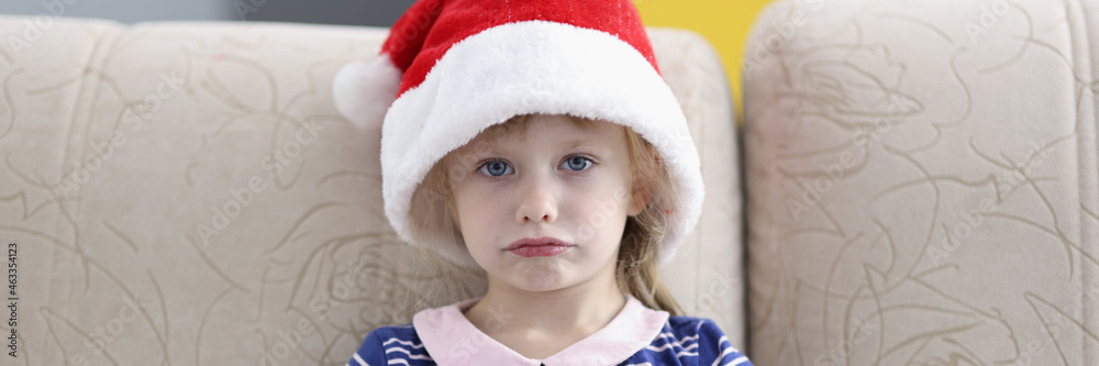 Little sad girl sitting on sofa wearing santa claus hat Little sad girl sitting on sofa wearing santa claus hat