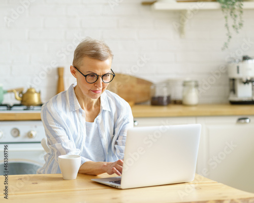 Modern focused mature woman writer wearing eyeglasses typing something on laptop, working online while sitting in kitchen at home. Senior people and technologies concept