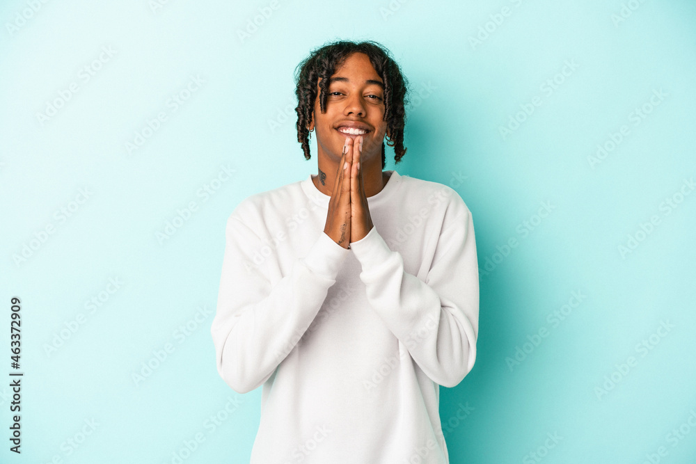 Young african american man isolated on blue background holding hands in pray near mouth, feels confident. Young african american man isolated on blue background holding hands in pray near mouth, feels confident.