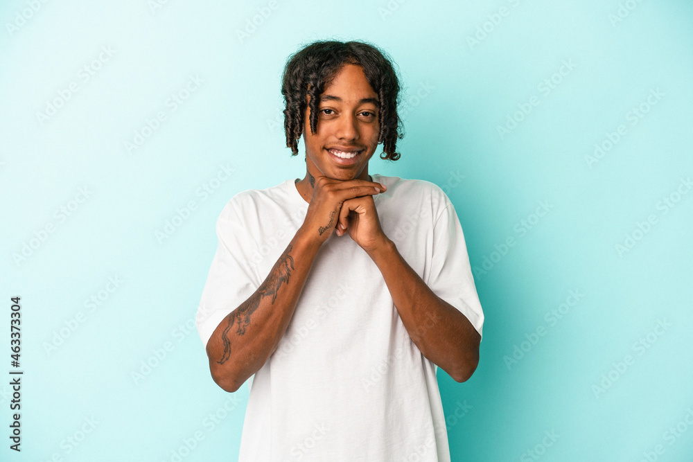 Young african american man isolated on blue background keeps hands under chin, is looking happily aside. Young african american man isolated on blue background keeps hands under chin, is looking happily aside.
