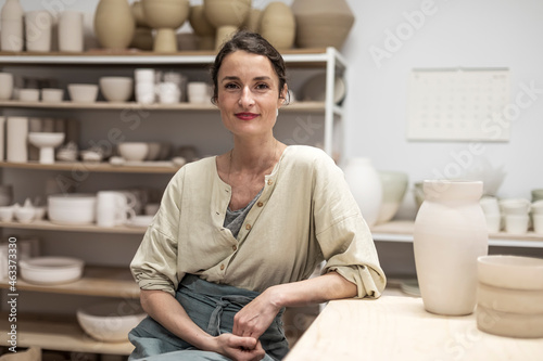 Portrait of beautiful happy craft woman wearing apron and smiling while sitting in her art studio or craft pottery shop