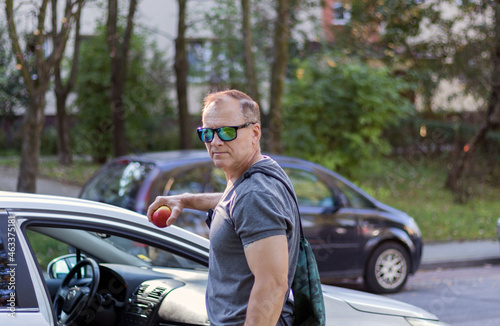 mature man, handsome courageous, 50 years old, dressed in a T-shirt, stands near the car and holds an apple, a man's portrait on a city background