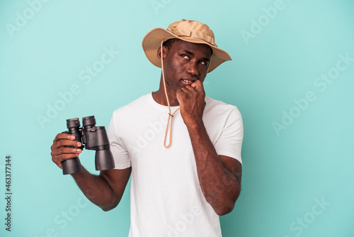 Young African American man holding binoculars isolated on blue background relaxed thinking about something looking at a copy space.