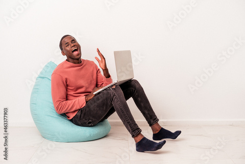 Young African American man sitting on a puff using laptop isolated on white background receiving a pleasant surprise, excited and raising hands.