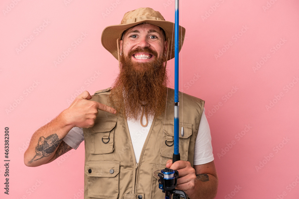 Young caucasian ginger fisherman with long beard holding a rod isolated on pink background person pointing by hand to a shirt copy space, proud and confident