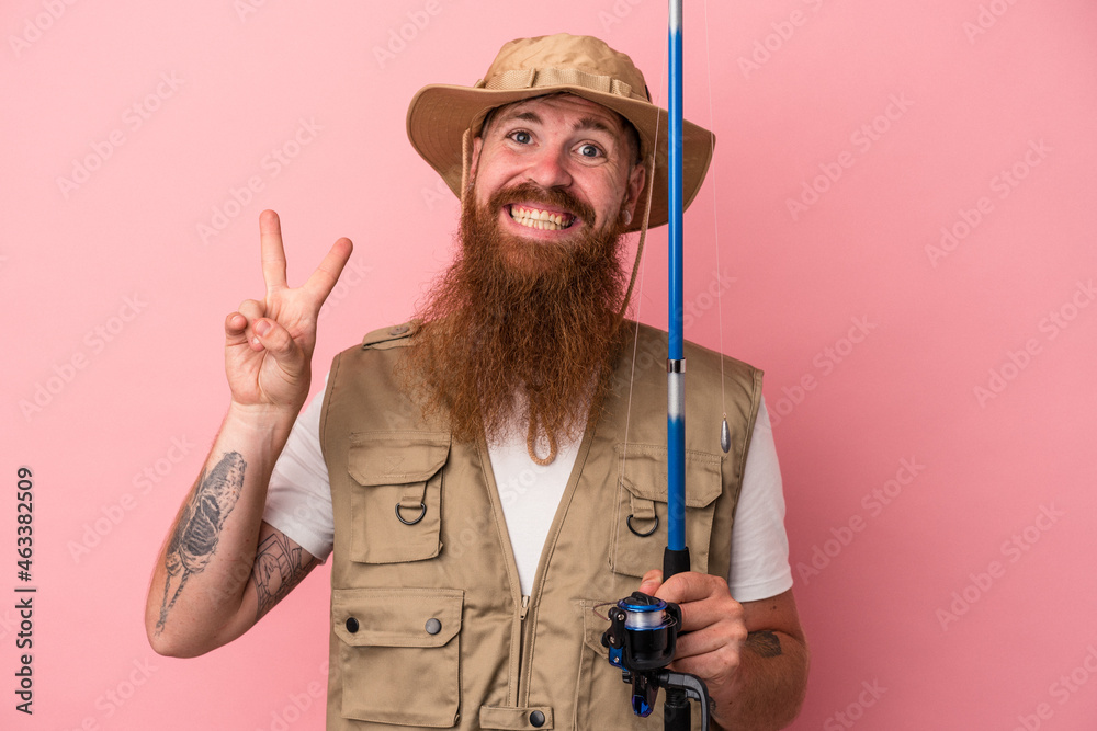 Young caucasian ginger fisherman with long beard holding a rod isolated on pink background joyful and carefree showing a peace symbol with fingers. Young caucasian ginger fisherman with long beard holding a rod isolated on pink background joyful and carefree showing a peace symbol with fingers.