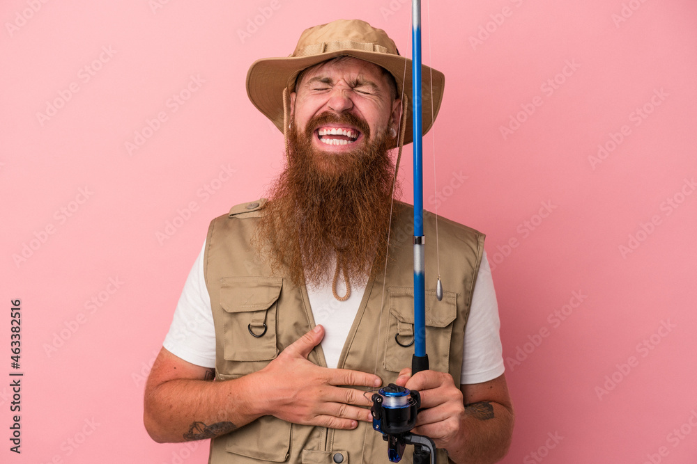 Young caucasian ginger fisherman with long beard holding a rod isolated on pink background laughing and having fun. Young caucasian ginger fisherman with long beard holding a rod isolated on pink background laughing and having fun.