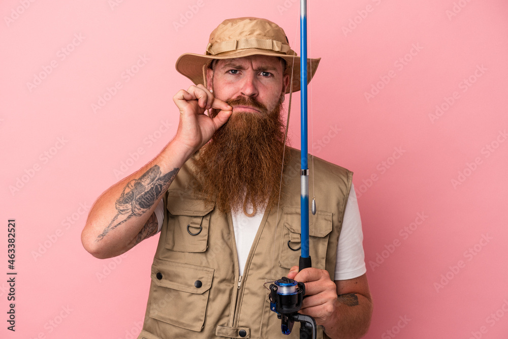 Young caucasian ginger fisherman with long beard holding a rod isolated on pink background with fingers on lips keeping a secret.