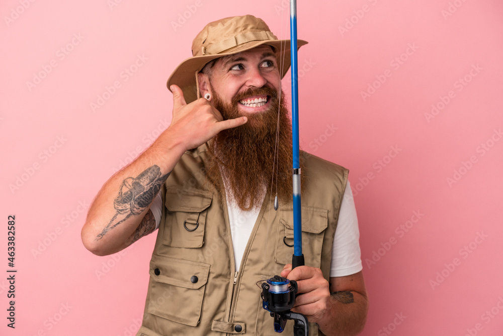 Young caucasian ginger fisherman with long beard holding a rod isolated on pink background showing a mobile phone call gesture with fingers. Young caucasian ginger fisherman with long beard holding a rod isolated on pink background showing a mobile phone call gesture with fingers.