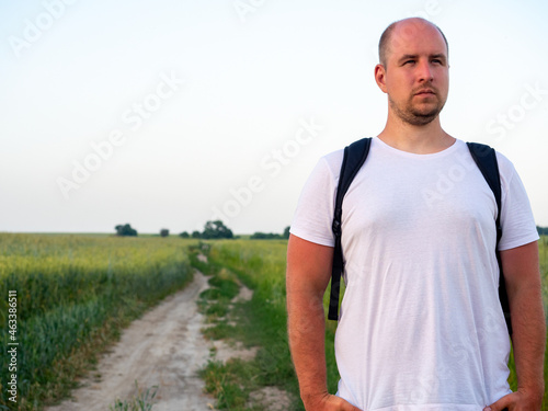 A man dressed in a T-shirt with a backpack on his back is standing in a field in the evening, . The path in the field in the background. The concept of travel. Copy space