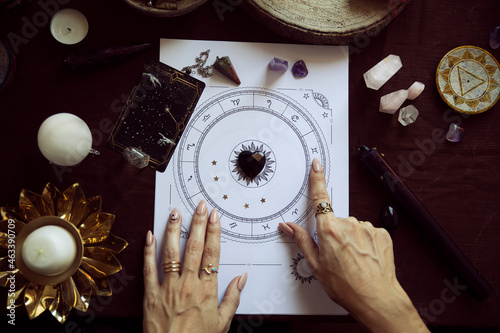 Top view of a woman pointing to a zodiac symbol in a person's birth chart on her witch altar with autumn colors, some candles and quartz crystals