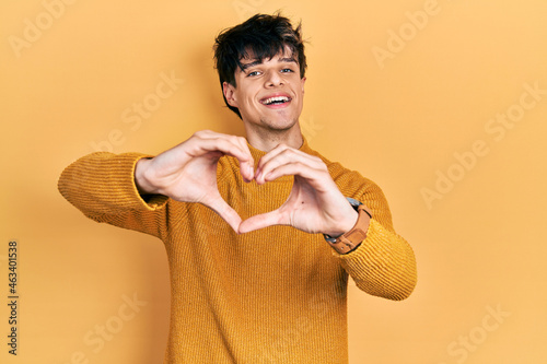 Handsome hipster young man wearing casual yellow sweater smiling in love doing heart symbol shape with hands. romantic concept.