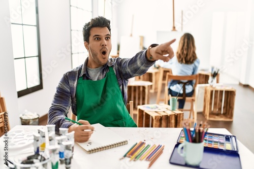 Young artist man at art studio pointing with finger surprised ahead, open mouth amazed expression, something on the front