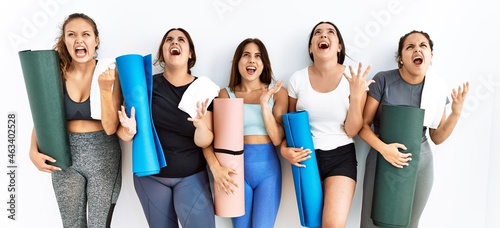Group of women holding yoga mat standing over isolated background crazy and mad shouting and yelling with aggressive expression and arms raised. frustration concept.