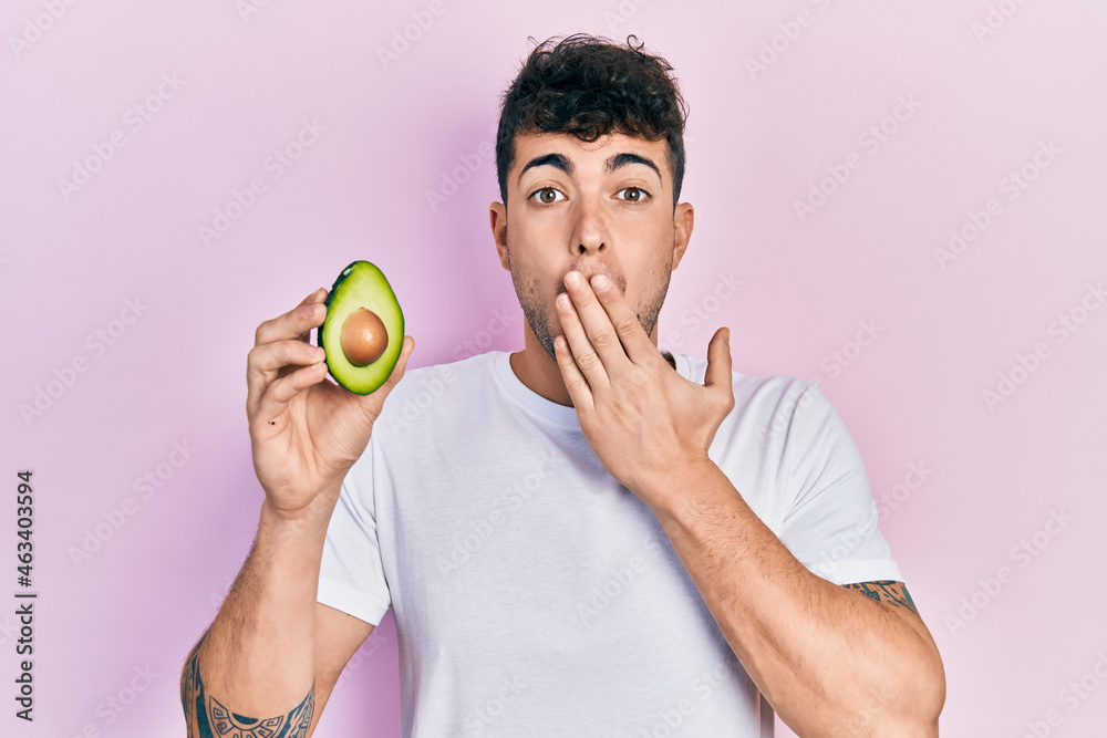 Young hispanic man holding middle avocado covering mouth with hand, shocked and afraid for mistake. surprised expression Young hispanic man holding middle avocado covering mouth with hand, shocked and afraid for mistake. surprised expression