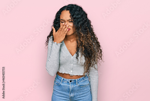 Young hispanic woman with curly hair wearing casual clothes smelling something stinky and disgusting, intolerable smell, holding breath with fingers on nose. bad smell