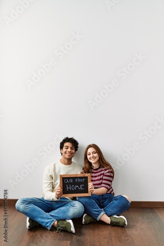 Young hispanic couple holding our first home blackboard sitting on the floor at empty new house.