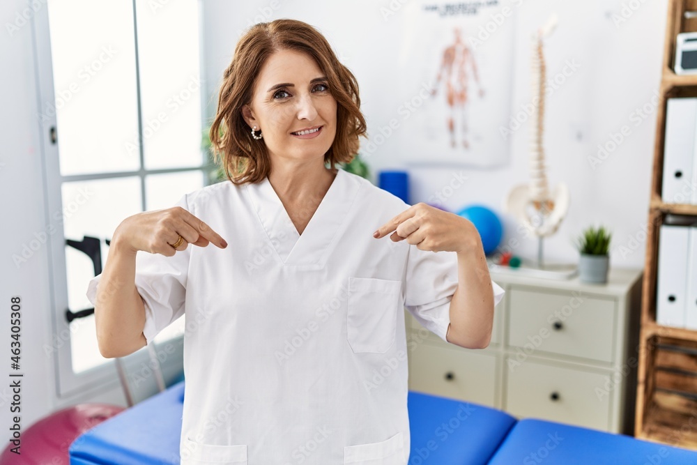 Fototapeta premium Middle age physiotherapist woman working at pain recovery clinic looking confident with smile on face, pointing oneself with fingers proud and happy.