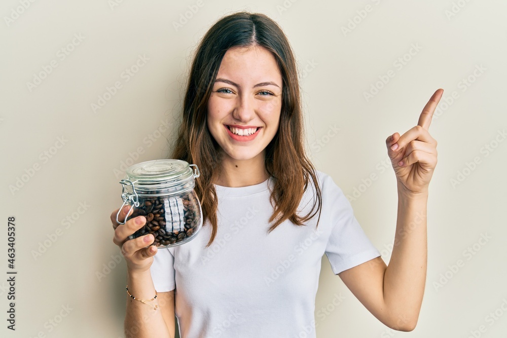 Young brunette woman holding jar with coffee beans smiling happy pointing with hand and finger to the side Young brunette woman holding jar with coffee beans smiling happy pointing with hand and finger to the side