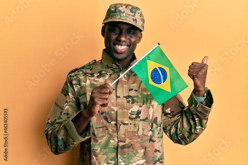 Young african american man wearing army uniform holding brazil flag pointing thumb up to the side smiling happy with open mouth