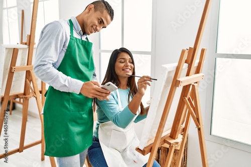 Young latin painter couple smiling happy painting and using smartphone at art studio