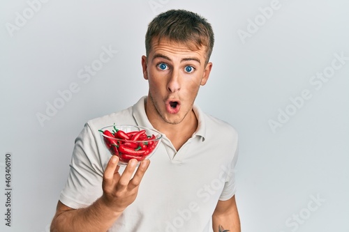 Young caucasian man holding bowl of red pepper scared and amazed with open mouth for surprise, disbelief face