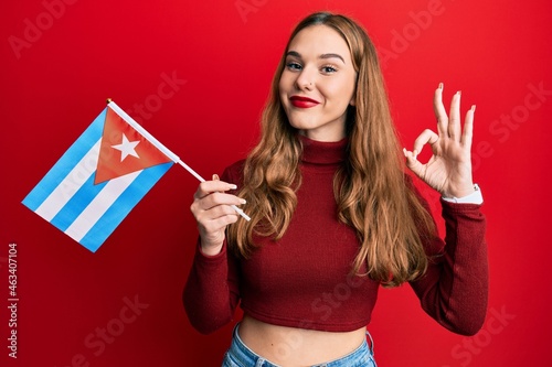 Photography Young blonde woman holding cuba flag doing ok sign with fingers, smiling friendl