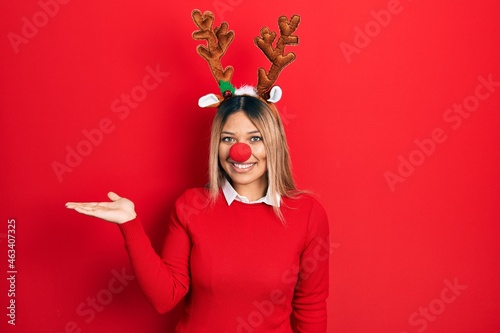 Beautiful hispanic woman wearing deer christmas hat and red nose smiling cheerful presenting and pointing with palm of hand looking at the camera.