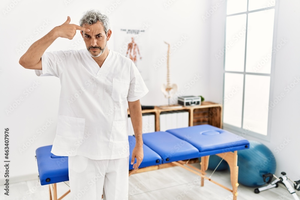 Obraz premium Middle age hispanic therapist man working at pain recovery clinic pointing unhappy to pimple on forehead, ugly infection of blackhead. acne and skin problem