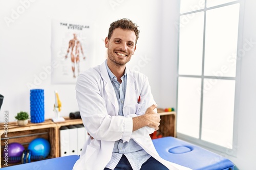 Young hispanic man wearing physiotherapist uniform with arms crossed gesture at clinic
