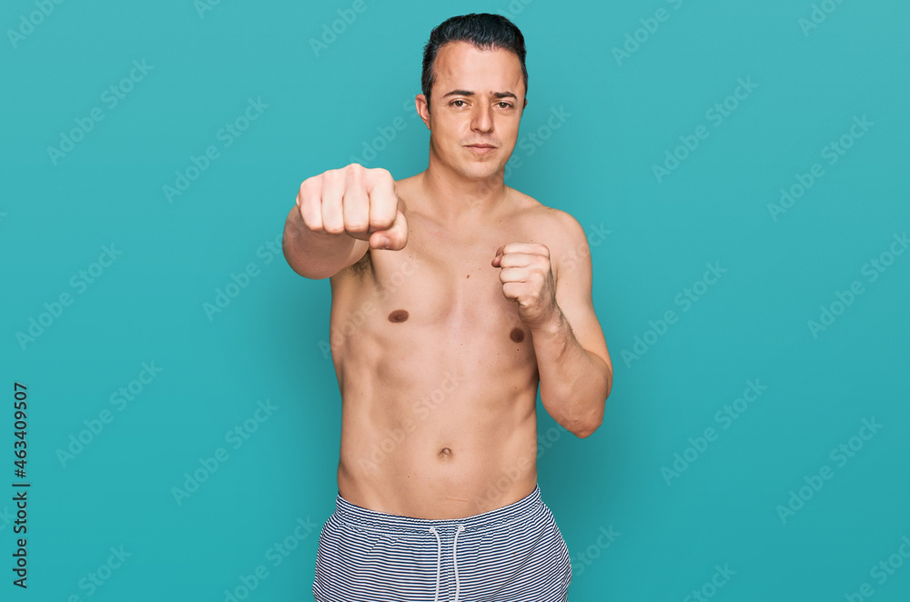 Fototapeta premium Handsome young man wearing swimwear shirtless punching fist to fight, aggressive and angry attack, threat and violence