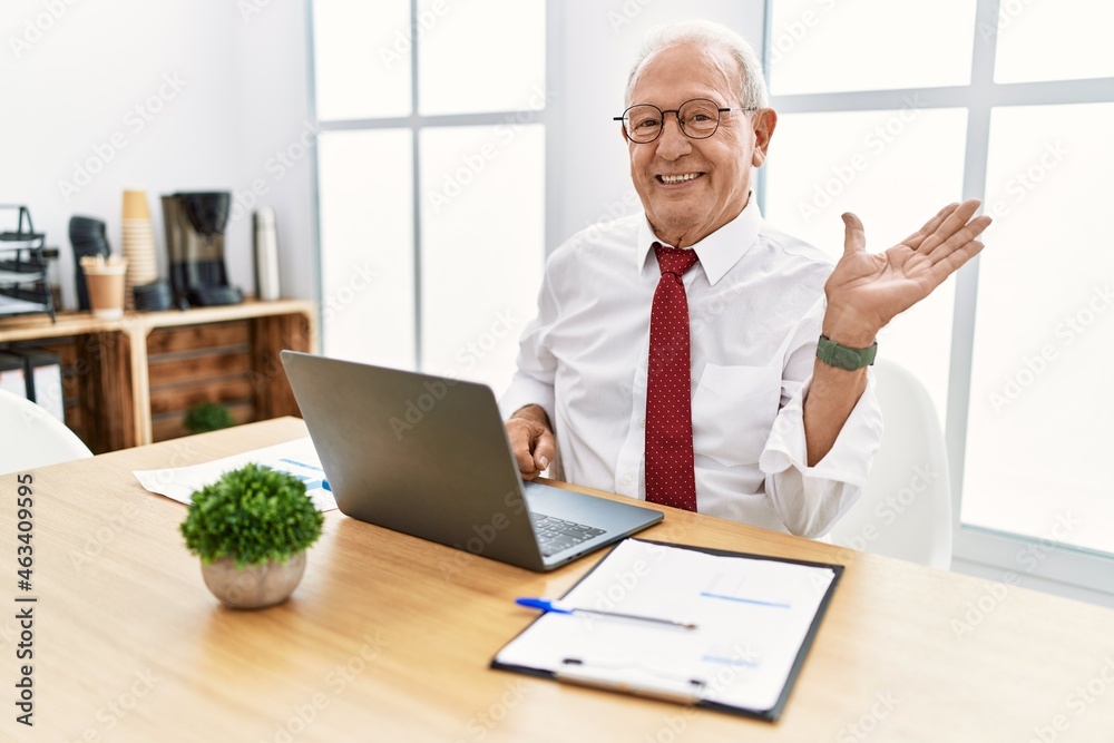 Senior man working at the office using computer laptop smiling cheerful presenting and pointing with palm of hand looking at the camera. Senior man working at the office using computer laptop smiling cheerful presenting and pointing with palm of hand looking at the camera.